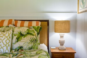 A bed with a tropical leaf pattern bedspread, a striped pillow, and a wooden nightstand with a lamp. - Waimea Beach Cottage by Destination Kauai - Charming 2BD, A/C, Walk to Ocean/Pier (Waimea)