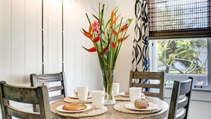 A round wooden table with white plates, two white cups, and a clear vase with red flowers. - Waimea Beach Cottage by Destination Kauai - Charming 2BD, A/C, Walk to Ocean/Pier (Waimea)