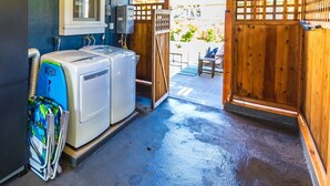 A laundry area with two white washers and dryers, a blue folding station, and a wooden gate leading to an outdoor patio. - Waimea Beach Cottage by Destination Kauai - Charming 2BD, A/C, Walk to Ocean/Pier (Waimea)