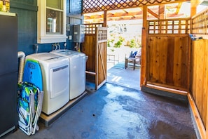 A laundry area with two white washers and dryers, a blue folding station, and a wooden gate leading to an outdoor patio. - Waimea Beach Cottage by Destination Kauai - Charming 2BD, A/C, Walk to Ocean/Pier (Waimea)
