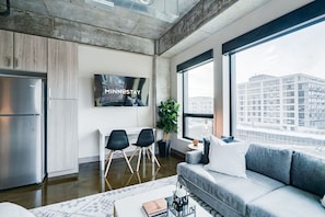A modern living room with a large window, a sofa, a TV, and a refrigerator. - MINNeSTAY*  Sable 301 - Studio | North Loop | Target Center (Minneapolis)
