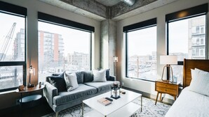 Moderni hotellihuone, jossa on harmaa sohva, valkoinen sohvapöytä, puinen yöpöytä ja sänky. Suurista ikkunoista avautuu näkymä kaupunkimaisemaan.