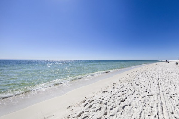 A wide sandy beach with footprints leading to the water, a clear blue sky, and a few people in the distance.