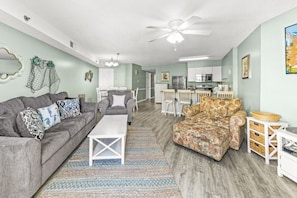 A living room with a grey sofa, a patterned armchair, a coffee table, and a kitchen in the background.