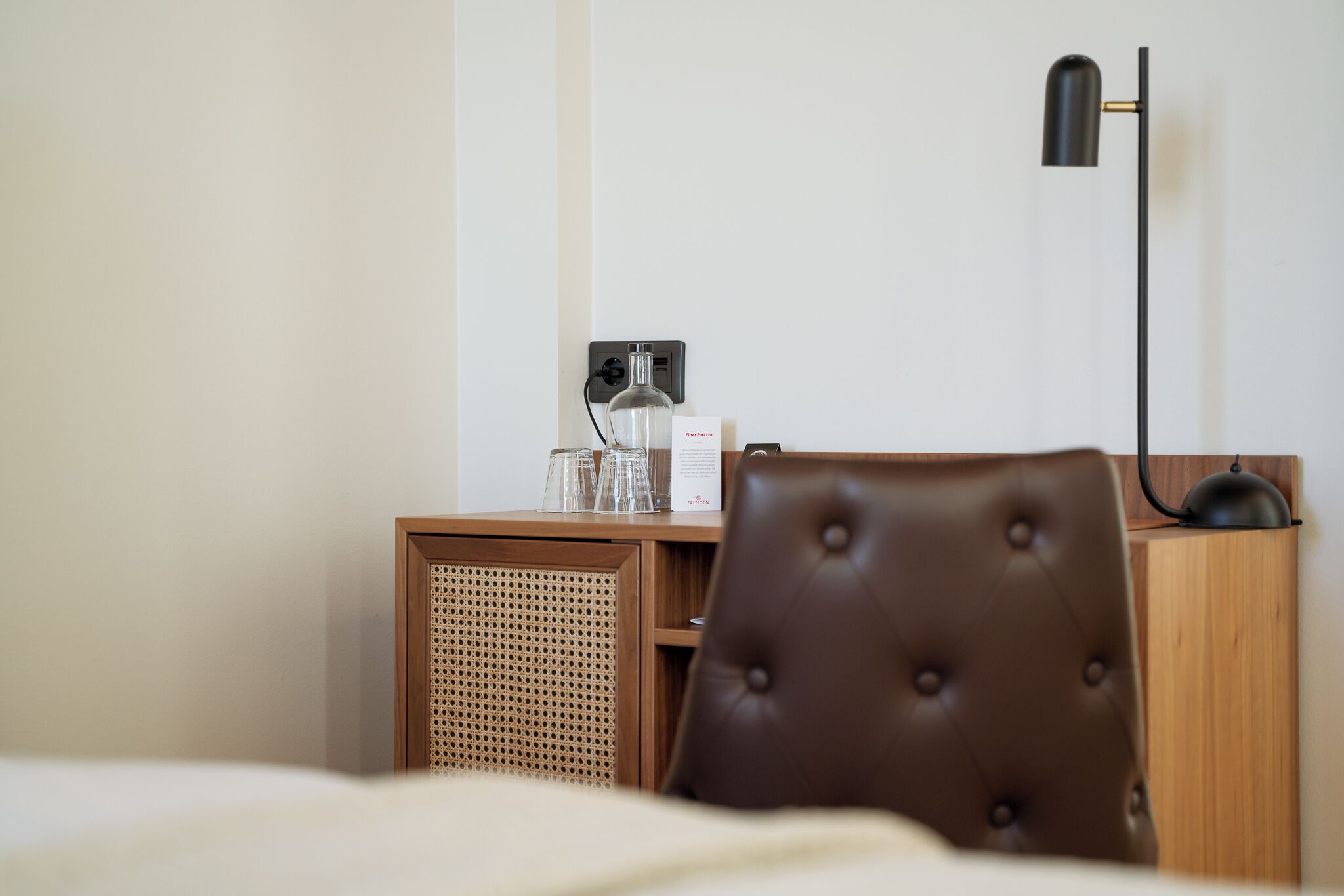 Hotellihuone, jossa on sänky, puinen y öpöytä lampun kanssa, tuoli ja pieni pöytä, jonka päällä on lasisetti.