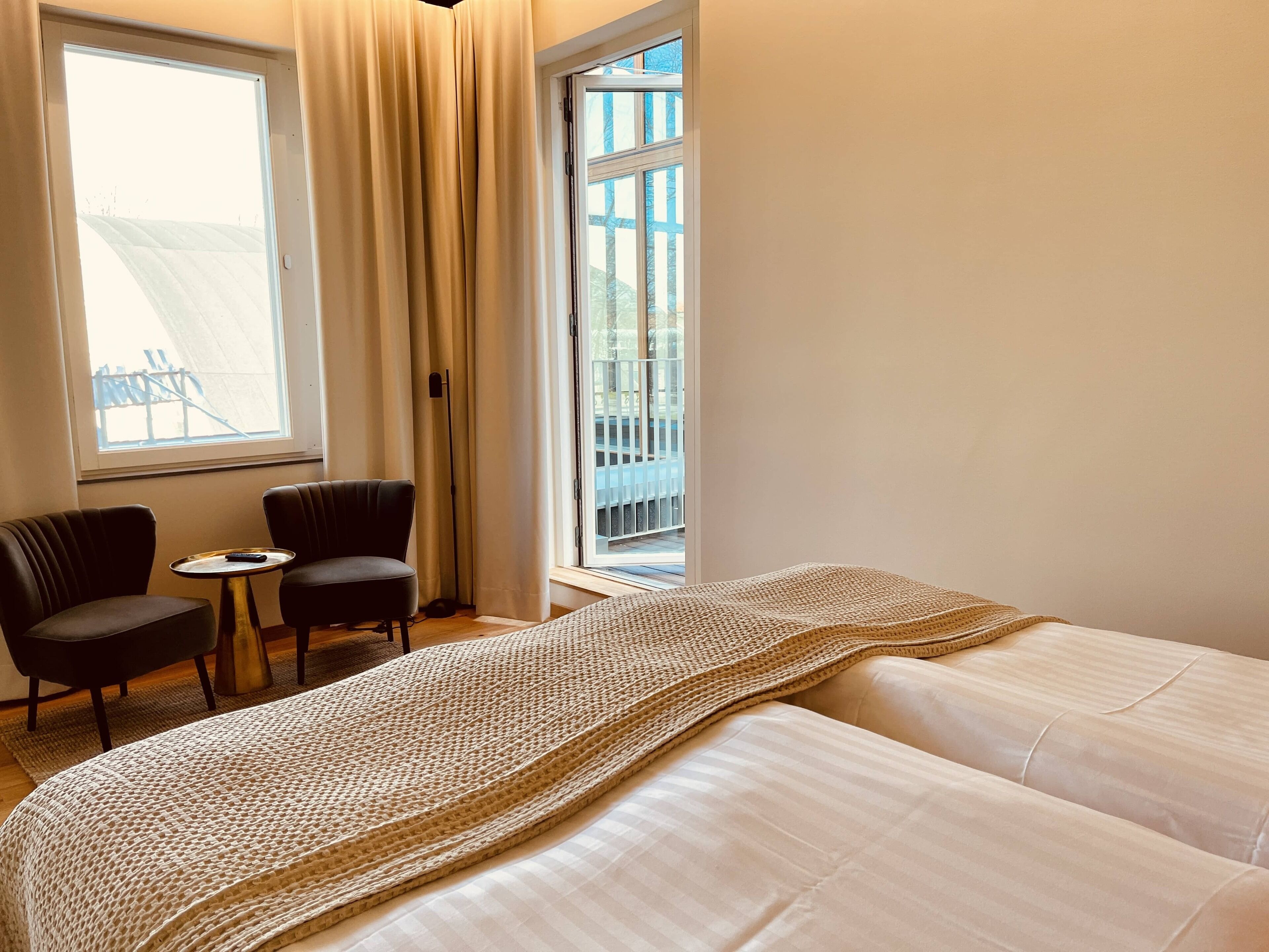 Ein Hotelzimmer mit einem Bett, zwei Stühlen, einem kleinen Tisch und einem großen Fenster mit Vorhängen.
