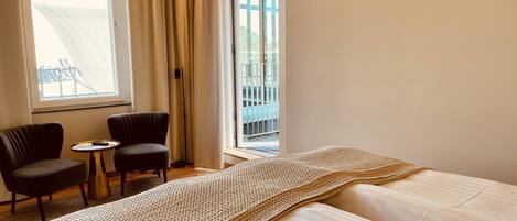 Een hotelkamer met een bed, twee stoelen, een kleine tafel en een groot raam met gordijnen.