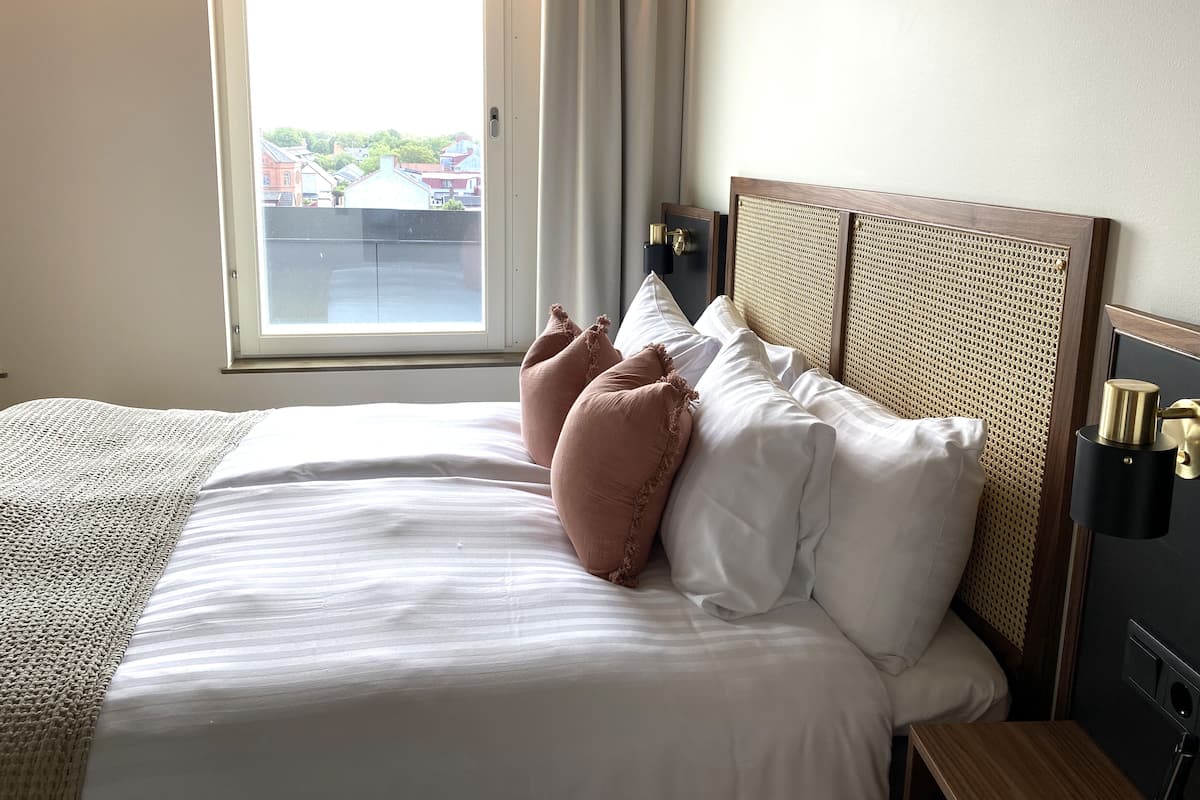 Een hotelkamer met een groot bed, een raam met gordijnen en een nachtkastje met een lamp.