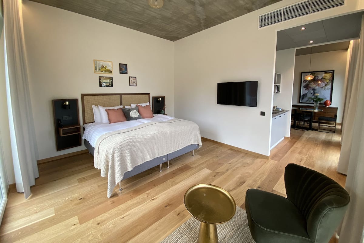 Een moderne hotelkamer met een houten vloer, een bed met een wit en grijs dekbedovertrek, een flatscreen-tv aan de muur en een zithoek met een groene fauteuil en een gouden kruk.