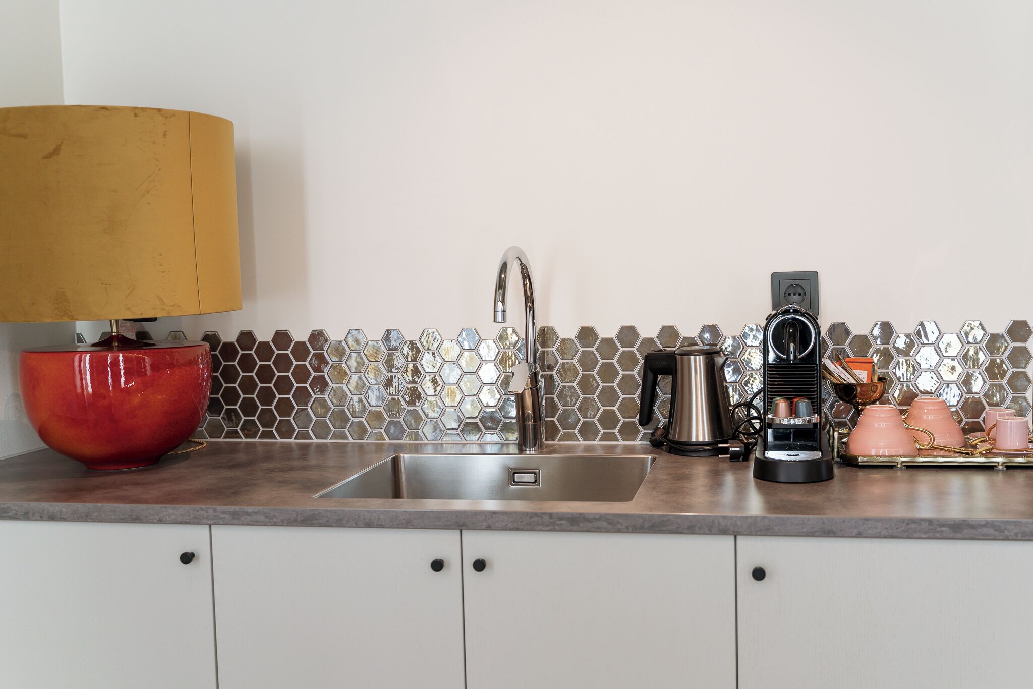 Een keuken met een spoelbak, een koffiezetapparaat, een rode kom en een zeshoekige spatwand.