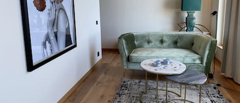 A living room with a green sofa, a small round table with two cups, a framed abstract painting, and a teal lamp.