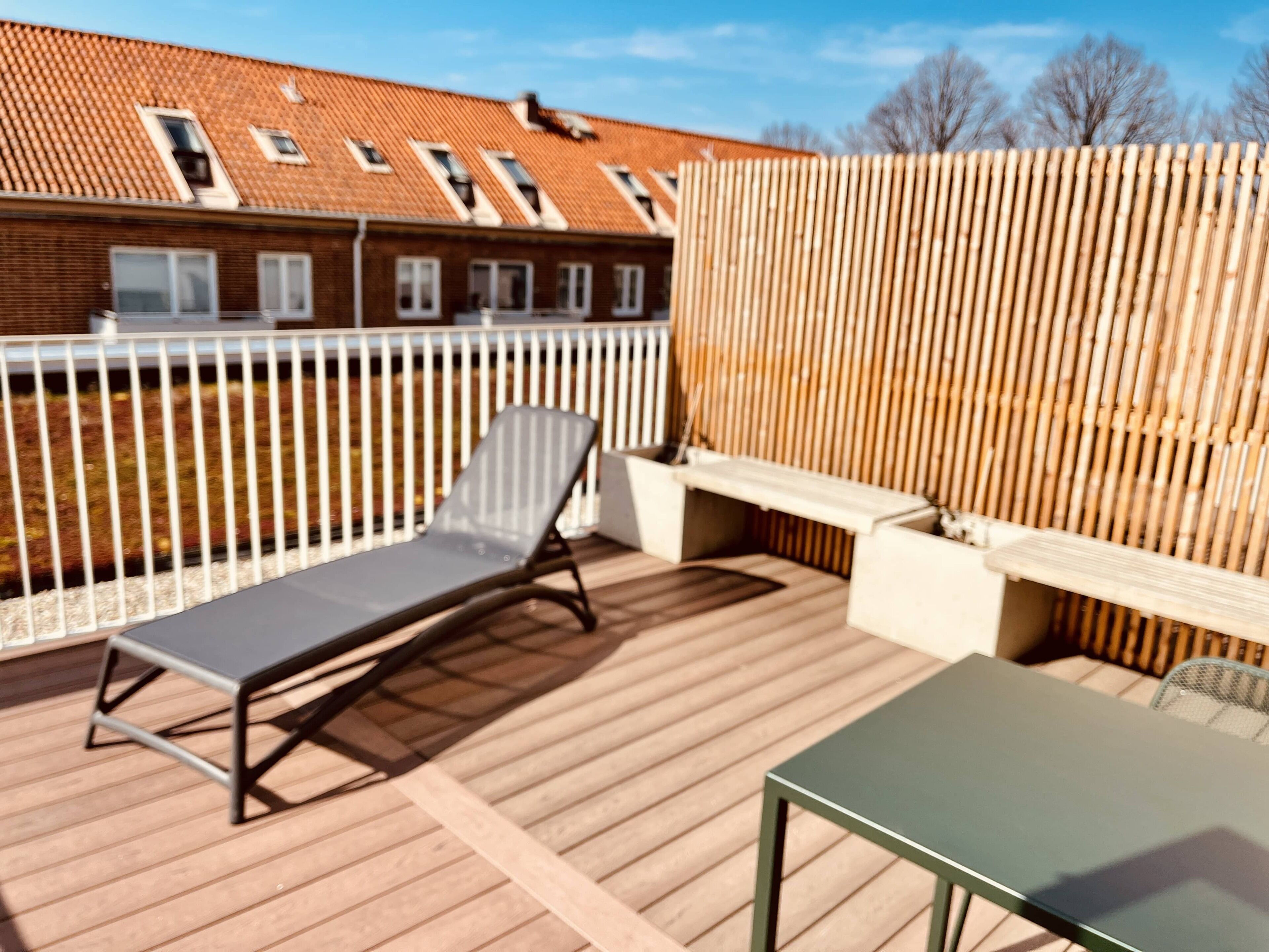 En träveranda med en solstol och ett bord, ett vitt staket och en byggnad i tegel med fönster i bakgrunden.