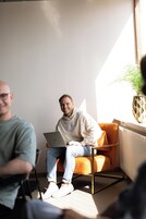 Une pièce dans laquelle une personne est assise sur une chaise orange et utilise un ordinateur portable. Une autre personne se trouve au premier plan, et une plante en pot est placée près de la fenêtre.