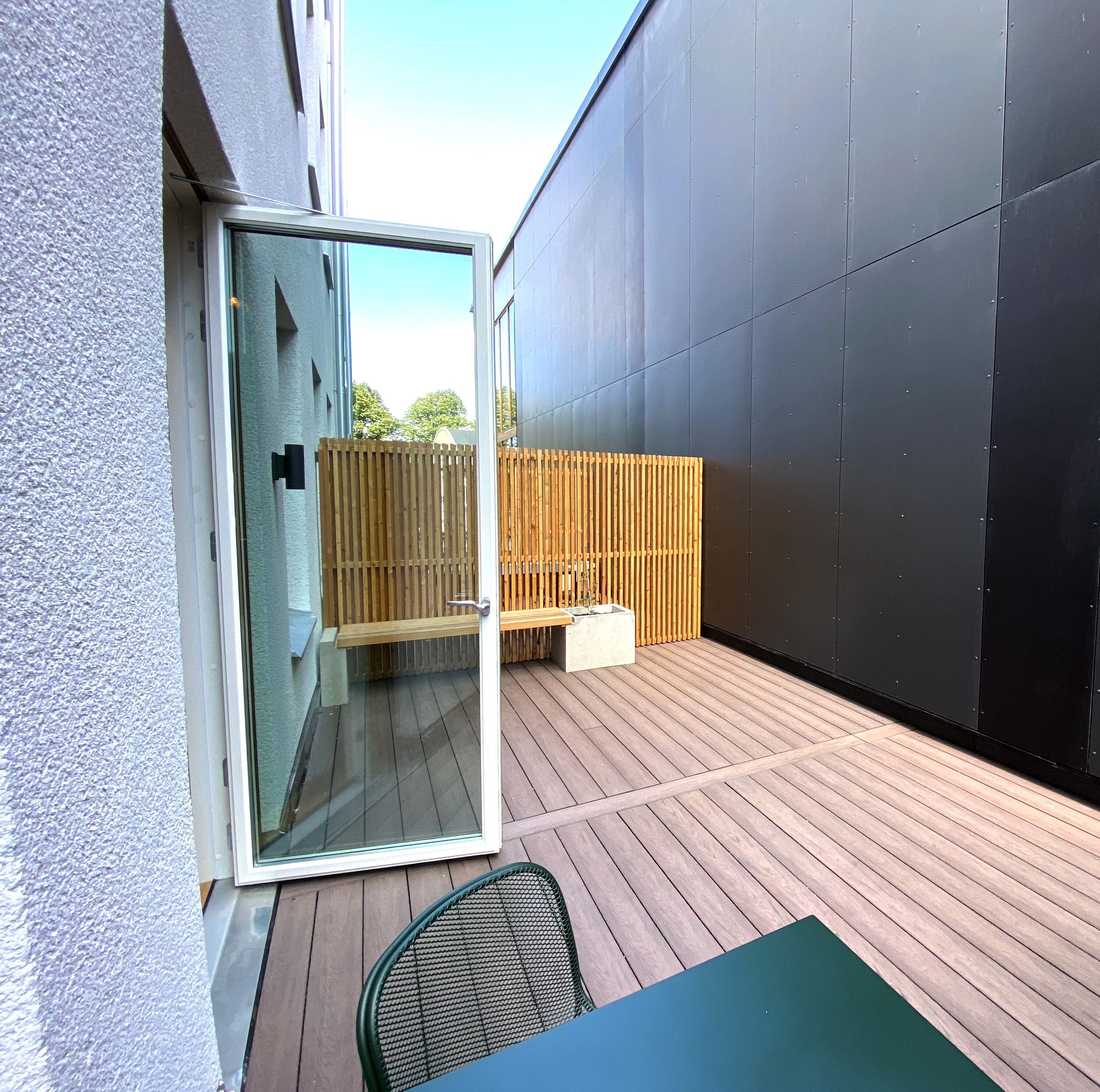 Una terraza de madera con un ventanal de vidrio que da a una habitación, un banco y una mesa con sillas.