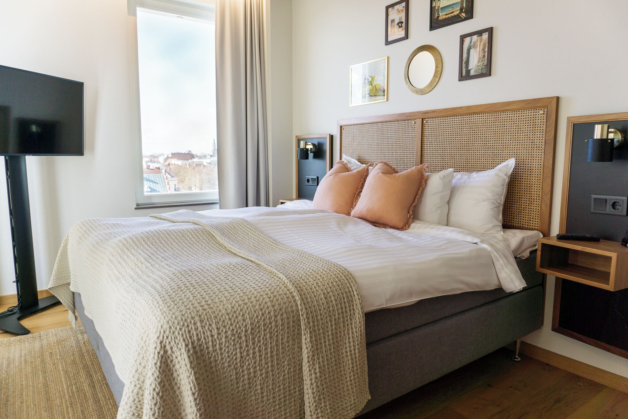 A neatly made bed with a grey mattress and beige bedding, a bedside table with a lamp, and a large window with curtains.