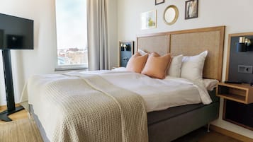 A neatly made bed with a grey mattress and beige bedding, a bedside table with a lamp, and a large window with curtains.