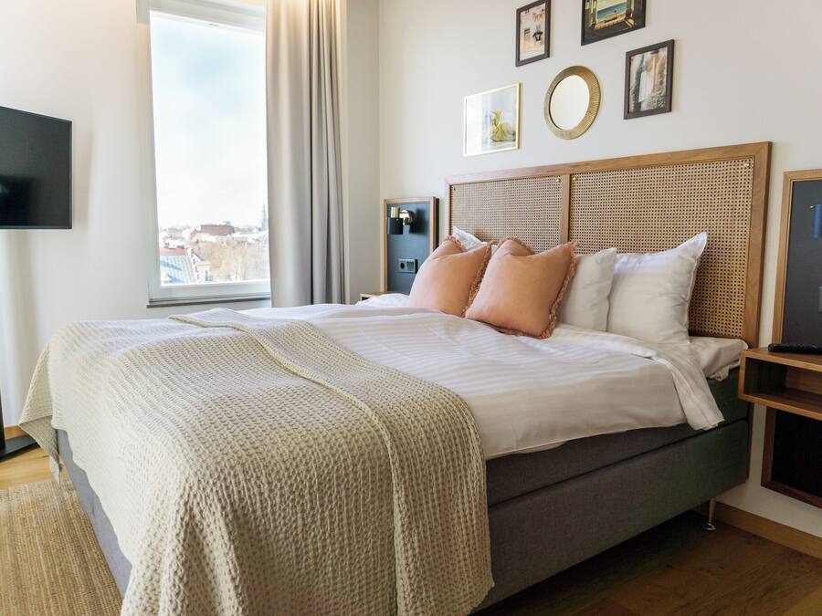 A neatly made bed with a grey mattress and beige bedding, a bedside table with a lamp, and a large window with curtains.