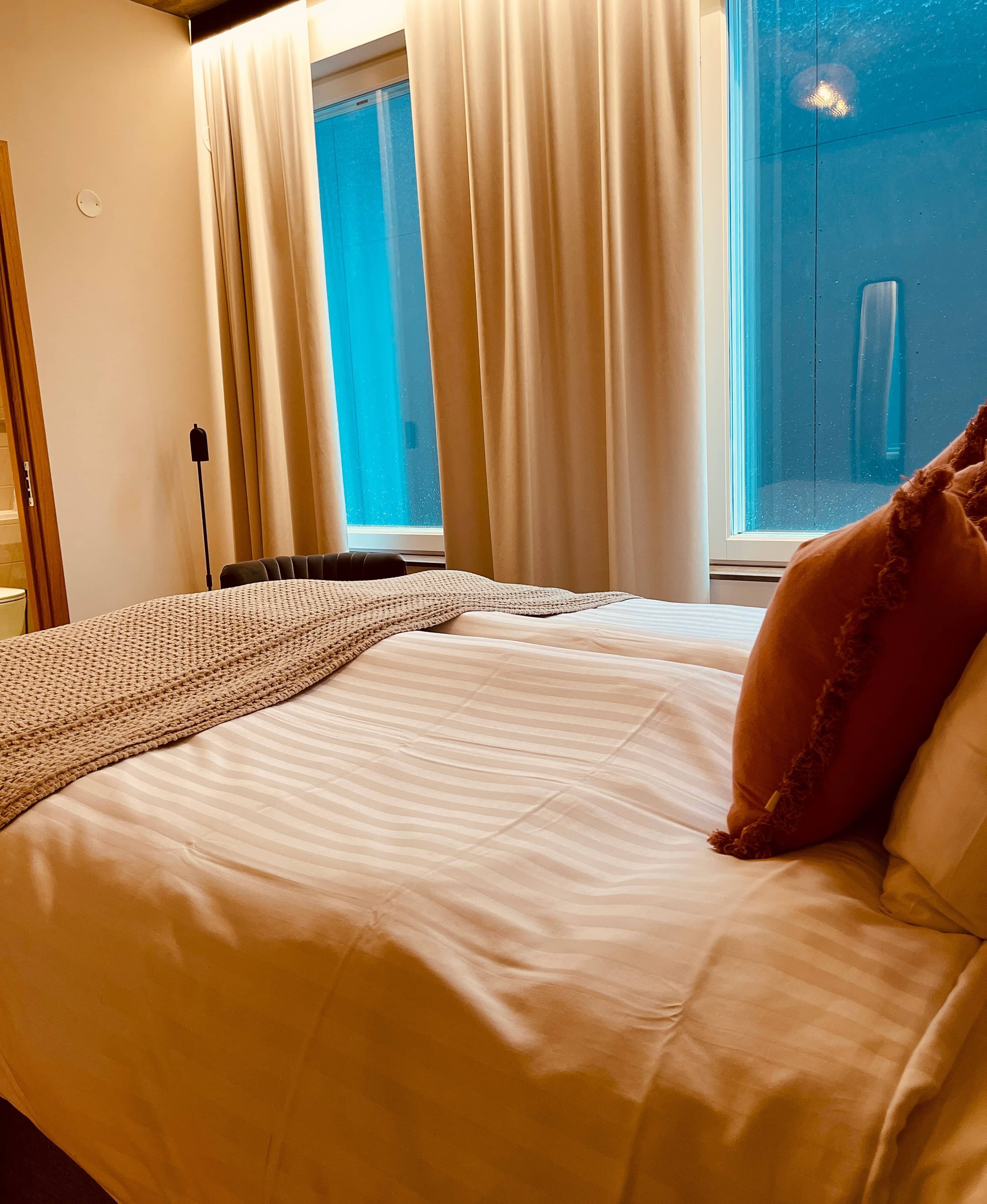 A neatly made bed with a striped blanket and pillows, a bedside lamp, and a window with curtains.