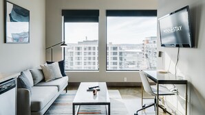 Una sala de estar moderna con un sofá, una mesa de centro y un escritorio con una computadora. Amplias ventanas ofrecen vista a la ciudad.