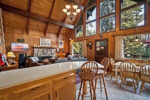Interior de una cabaña de madera con una gran mesa de comedor, sillas, chimenea y zona de cocina.
