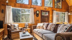 Interior acolhedor de uma cabana com paredes de madeira, um sofá, uma poltrona, um ventilador e um relógio na parede.
