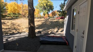 A covered porch with a concrete floor, a door, and a small step leading to a gravel area with trees and a rock. - Modern Studio excellent location. (Grand Junction)