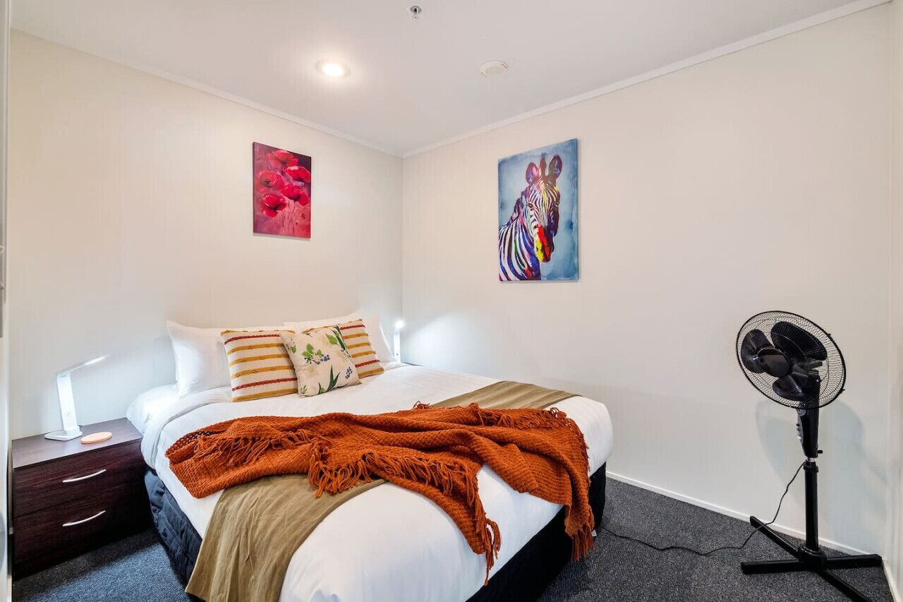 A bedroom with a bed, pillows, a blanket, a nightstand, a lamp, and a wall-mounted fan.