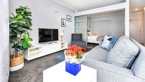 En moderne stue med en grå sofa, et hvidt sofabord og et fjernsyn på et hvidt skab. Der er en stor, grøn plante og en blå vase med blomster på sofabordet.
