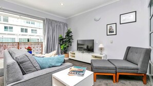 En moderne stue med en grå sofa, en grå lænestol, et hvidt sofabord og et fjernsyn på et hvidt skab. Der er et vindue med gardiner, en potteplante og billeder i rammer på væggen.