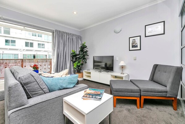 A modern living room with a grey sofa, a grey armchair, a white coffee table, and a TV on a white cabinet. There is a window with curtains, a potted plant, and framed pictures on the wall.