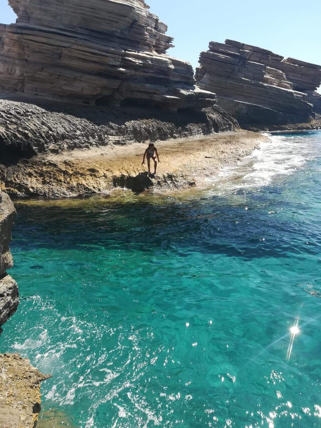 A rocky coastline with clear turquoise water and a person standing on the rocks.