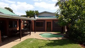 A backyard with a swimming pool, a covered patio with outdoor furniture, and a house with a green roof.