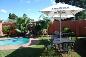 A backyard with a swimming pool, a stone wall, and a table with chairs under a branded umbrella. - D & H Guesthouse (Graskop)