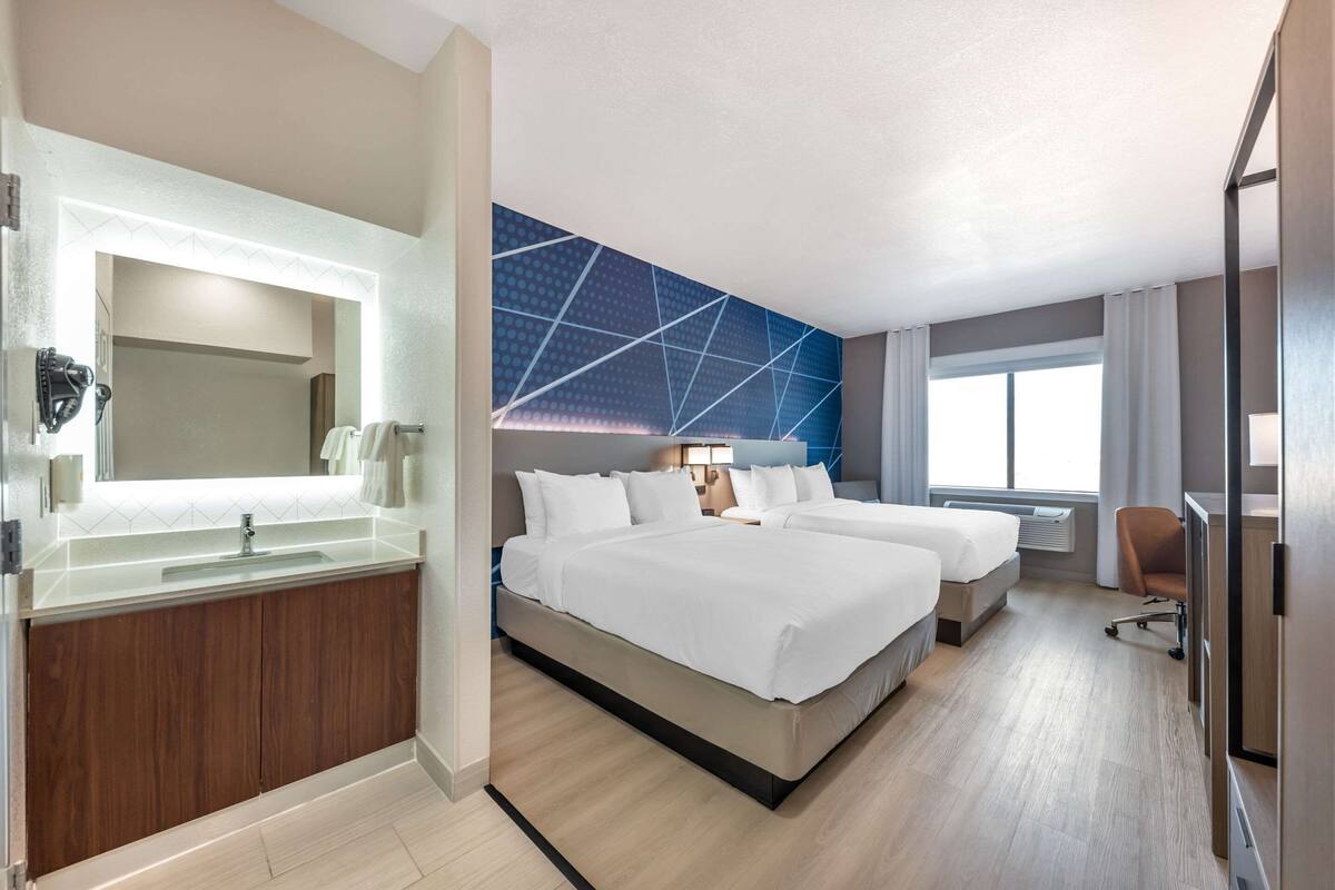 Une chambre d’hôtel avec un grand lit, un lit séparé, une salle de bain avec un miroir et un lavabo, et une vue sur l’extérieur.