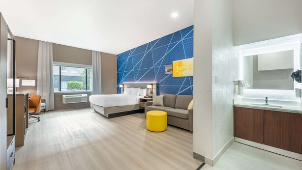 A modern hotel room with a bed, a sofa, a yellow ottoman, a desk, and a bathroom with a mirror and sink. - Comfort Inn & Suites Belton Temple South I-35 (Belton)