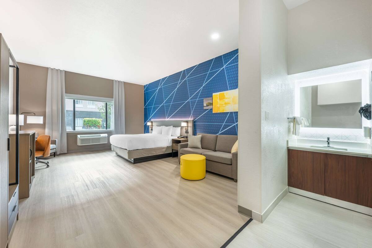 Une chambre d’hôtel moderne comprenant un lit, un canapé, un pouf jaune, un bureau et une salle de bain avec un miroir et un lavabo.