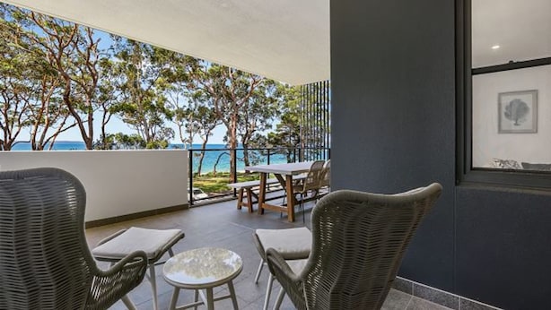 A balcony with wicker furniture, a table, and chairs, overlooking a coastal area with trees and the sea. - 204 on the Beach by Experience Jervis Bay (Huskisson)