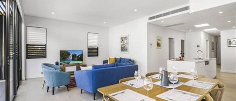 A modern living room with a blue sofa, a dining table set for two, and a kitchen area in the background.