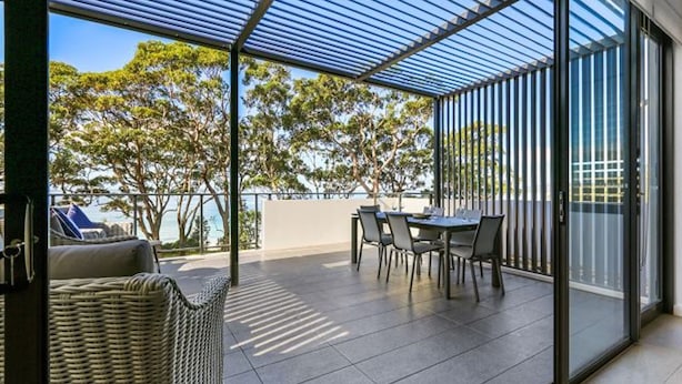 A covered outdoor area with a table and chairs, a wicker chair, and a view of trees and a body of water.