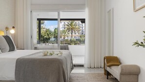 Ett hotellrum med en säng, en liten soffa, en balkong med utsikt över palmer och byggnader samt ett fönster med gardiner.