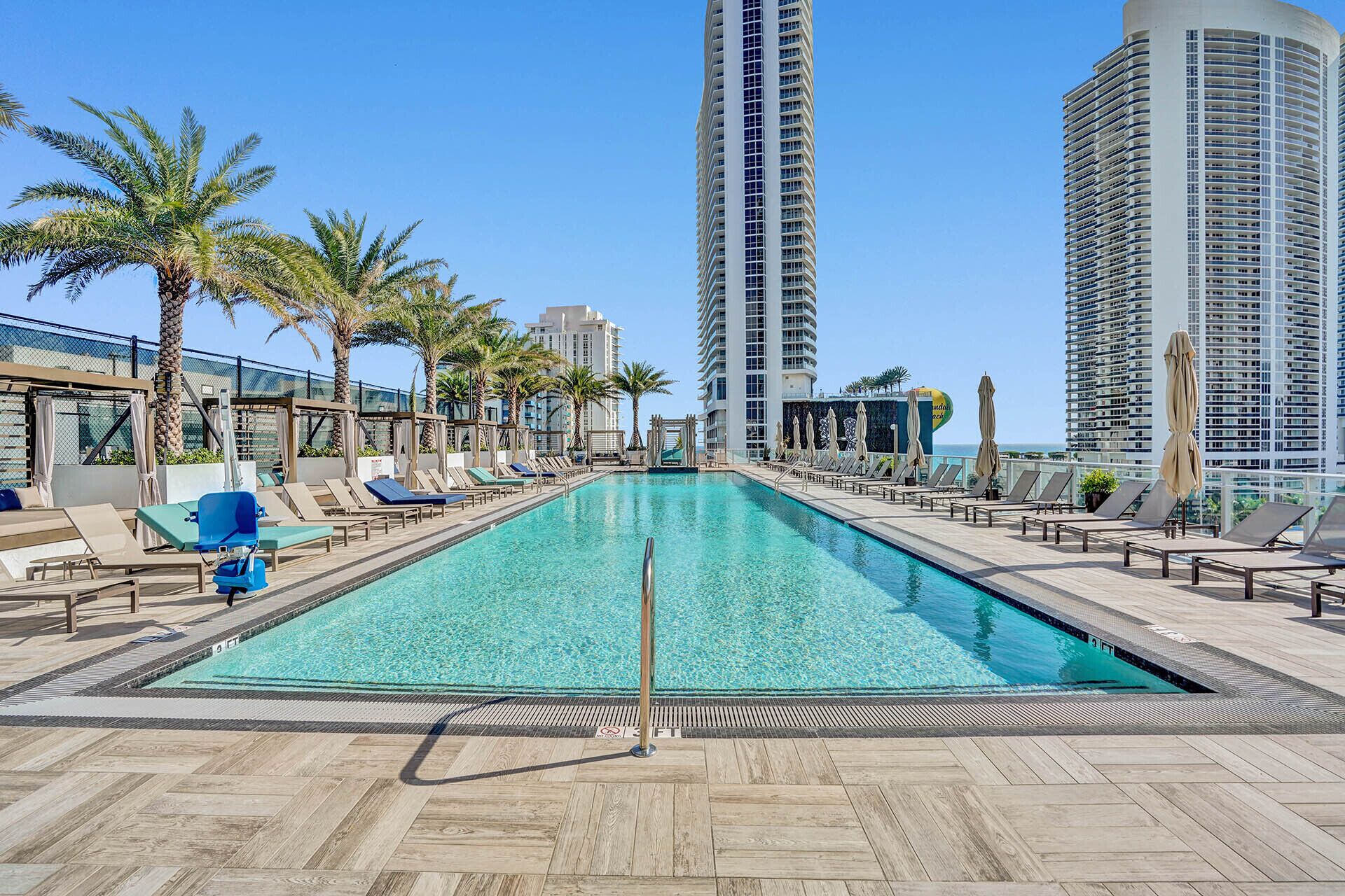 A rooftop pool with lounge chairs and palm trees, surrounded by high-rise buildings.