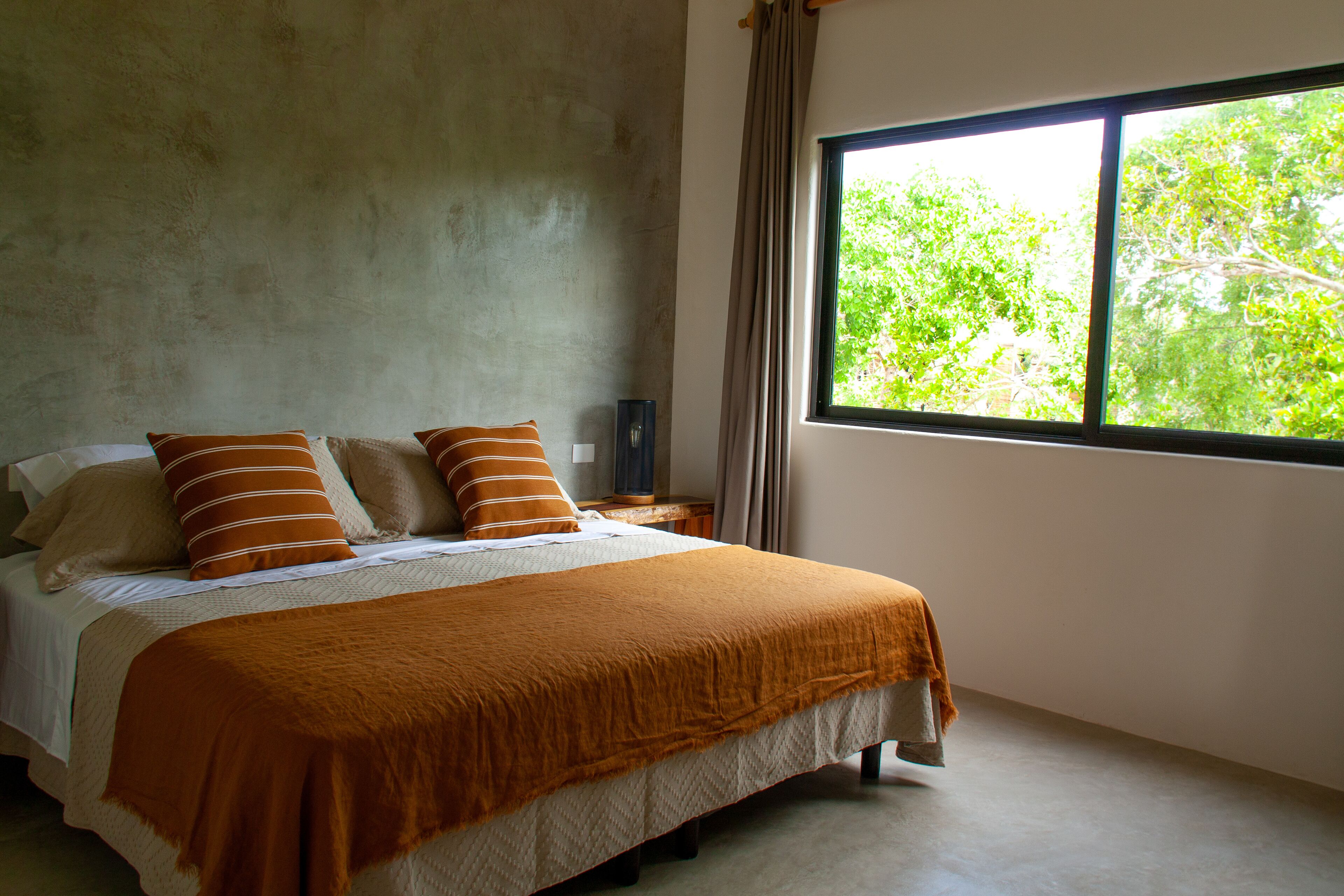 A bedroom with a large window, a bed with a brown blanket, and a concrete wall.