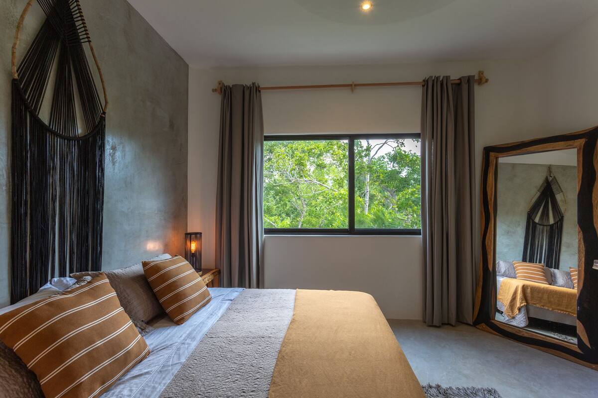 A bedroom with a large bed, a window with curtains, a wall-mounted macrame piece, and a mirror with a decorative frame.