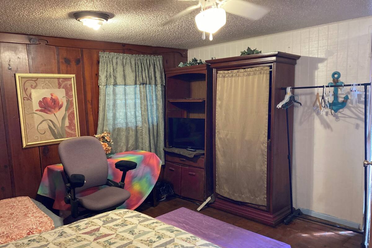 A room with a bed, a chair, a table, a wardrobe, a television, and a curtain.