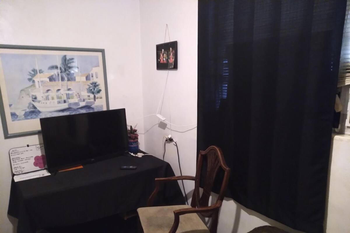 A room with a desk, a chair, a TV, a small table, a picture on the wall, and a window with curtains.