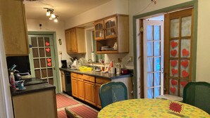 Una cocina con armarios de madera, una mesa de comedor con mantel estampado de frutas y una ventana decorada con corazones.