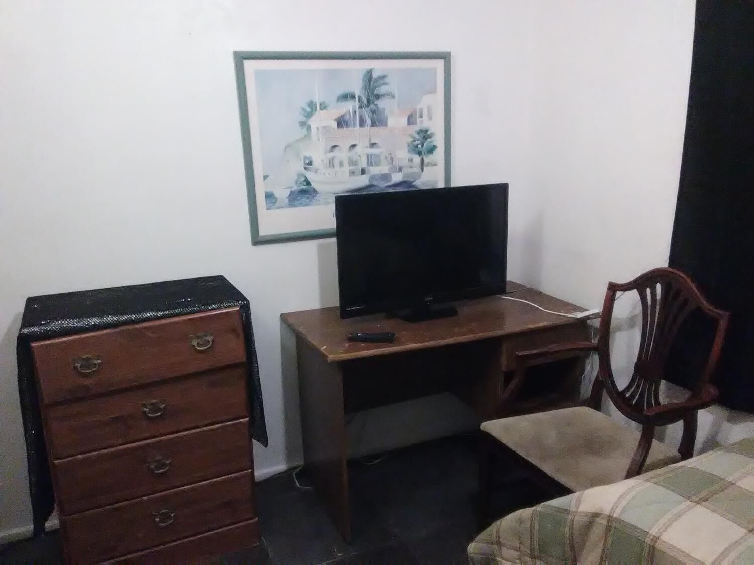Ein kleines Zimmer mit einem Holztisch, einem Fernseher, einem Sessel, einer Kommode und einem Bett mit karierter Decke.