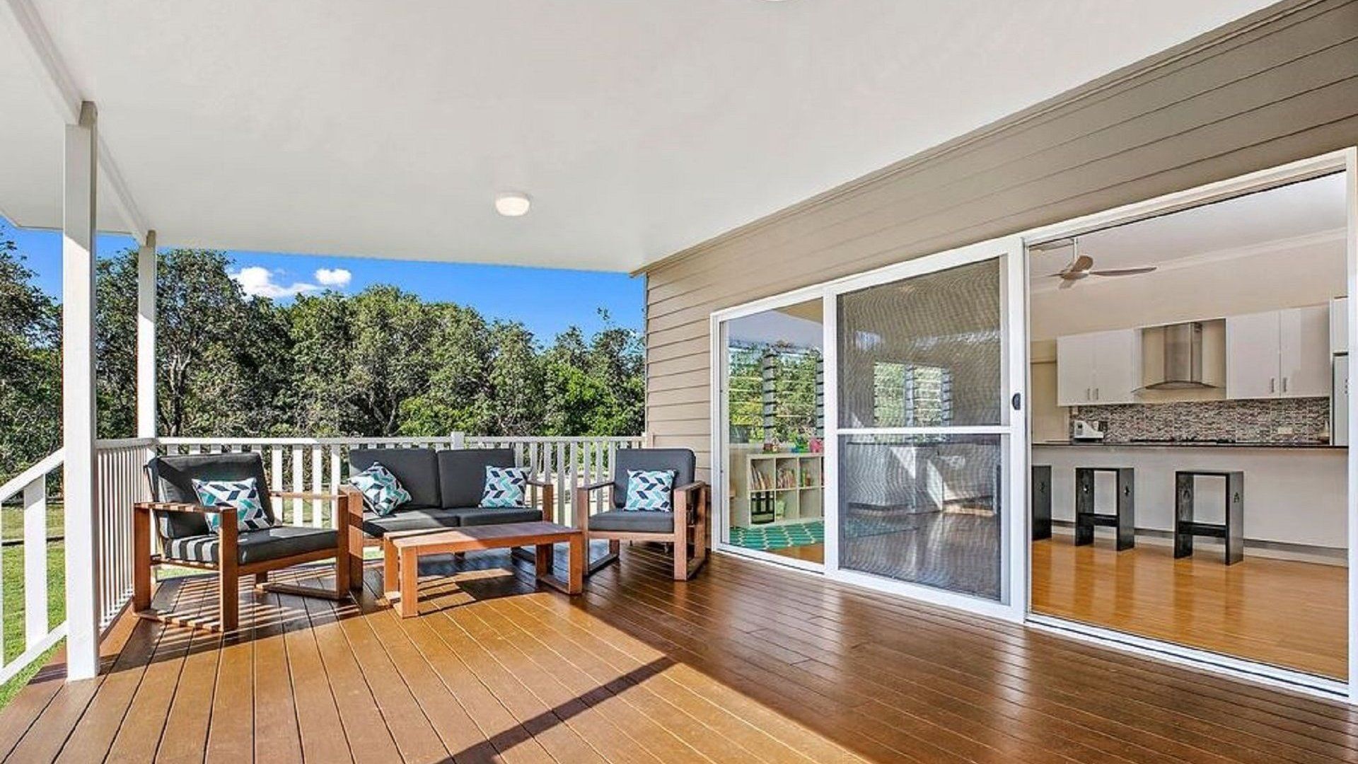 A covered outdoor deck with wooden flooring, a sofa set, and a sliding glass door leading to a kitchen area.