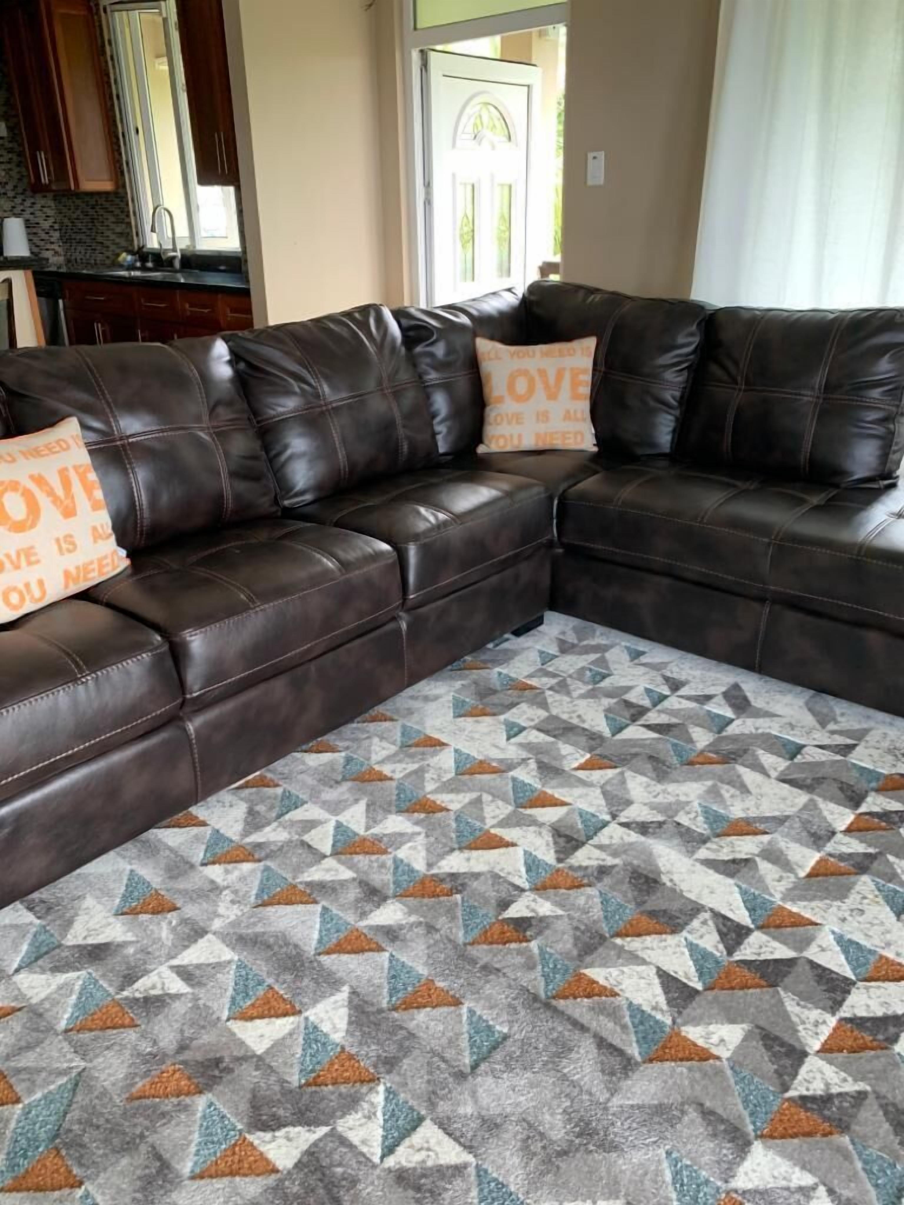 Ein braunes, ledernes Ecksofa mit zwei Kissen, einem gemusterten Teppich und einer Küche im Hintergrund.