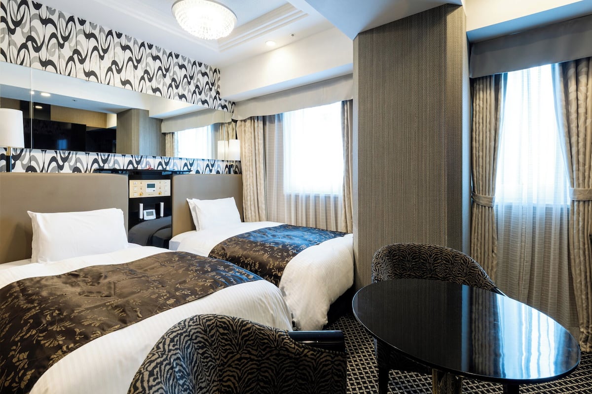 A hotel room with two beds, a desk, a chair, and a large window with curtains.